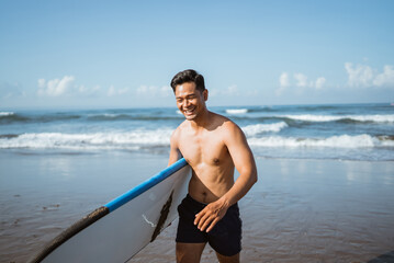 Asian male surfer happily carrying a surfboard after training during a beach walk