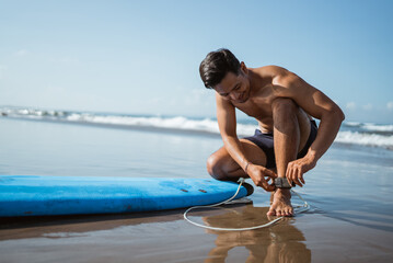 man attaches a safety harness to connect his feet to a surfboard before surfing at the beach - Powered by Adobe