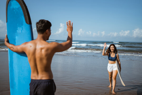 female surfer greets a man with a wave on the beach with her surfboard after practicing