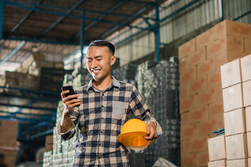 asian man factory employee removes safety helmet while using cell phone in warehouse - obrazy, fototapety, plakaty