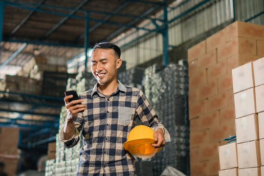 asian man factory employee removes safety helmet while using cell phone in warehouse