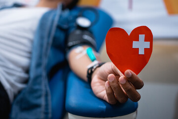 male donor hand lying holding heart shape paper with white cross during blood transfusion in...