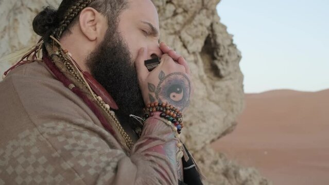 Man dressed as a shaman, standing on a rock in the middle of the desert sands and playing the harmonica