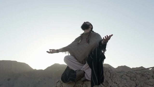 A youthful young shaman in the lotus position prays on a rock against the backdrop of sunset among the sand dunes of the desert