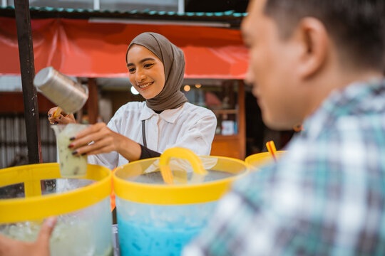 a pretty girl in a veil sells es campur using a scoop to get coconut milk from a jar on a cart