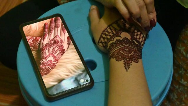 Artist applying henna tattoo on women hands while looking at pictures on a smartphone.