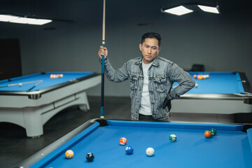 Fototapeta premium asian pool player put his hand on his waist while standing with sullen face and holding the cue stick
