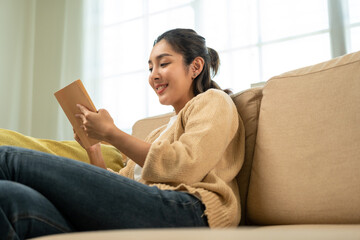 Asian beautiful woman sitting reading book on cozey couch sofa in living room with sun light morning. People female stay home part time relax at home.