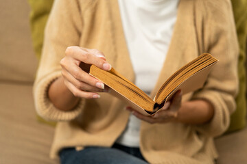 Close up female hands holding book turning the page. Young woman sitting reading book on cozey couch sofa in living room with sun light morning. People female stay home part time relax at home.