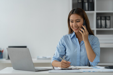 Happy Asian businesswoman talking on the phone and smiling while sitting at work using smartphone to talk. Contact via online application, concept of management