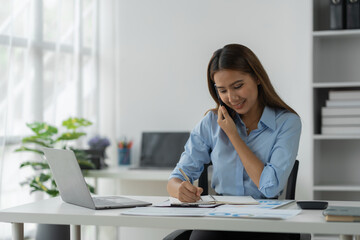 Happy Asian businesswoman talking on the phone and smiling while sitting at work using smartphone to talk. Contact via online application, concept of management