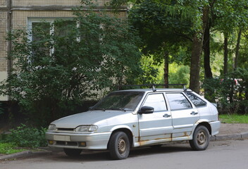An old rusty silver car is parked near a residential building, 