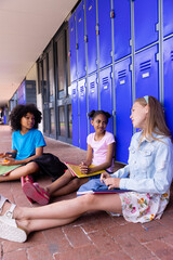 Happy diverse schoolchildren sitting next to lockers in corridor at school