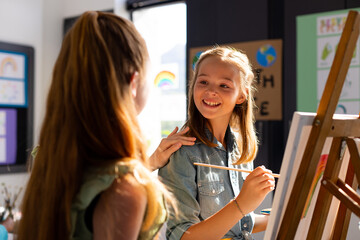 Fototapeta premium Happy diverse schoolchildren painting using brush and easel in school art class
