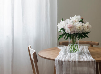 A bouquet of beautiful white peonies in a glass vase on a wooden round table with a white tablecloth
