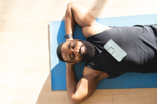 Portrait of relaxed african american man wearing sportswear with smartphone and lying on mat at home