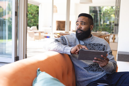 Thoughtful african american man in jumper, using tablet and looking through window on couch at home