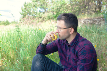 A young man sits near the forest and eats an apple. Rest in the open air. It's nice to relax in the park