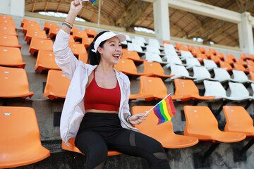 Happy and excited young female asian transgender LGBT sport fan cheering and watching the match with rainbow flag raising and wristband, she sitting amongst the rows of empty seats on stadium