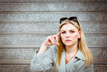 Portrait of a young blonde woman in a coat with sunglasses in her hair waiting on a staircase.
