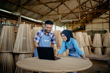 two smiling Asian entrepreneurs using laptop computer together on wooden table at a woodcraft shop
