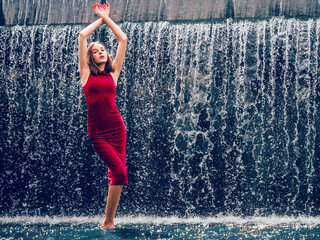 Teen ager girl in high fashion red dress posing in water, water fall behind her back. Prom photoshoot in beautiful location Young lady model ready for prom party. Confident person. Selective focus. © mark_gusev