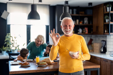 Happy confident senior man looking at camera and smiling showing with hand ok sign satisfied with breakfast porridge, standing in front of family members, senior woman and two kids in the kitchen.