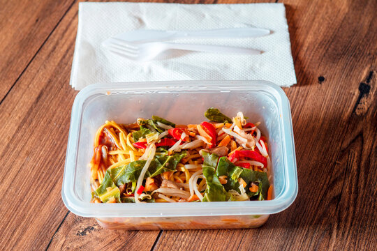 Chicken chow mein noodles in a plastic take away box or container on a wooden table. Quick and tasty Asian style meal for microwave. Lunch in an office concept. Food on a go. High quality ready meal.