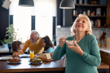 Happy satisfied senior woman enjoying porridge for breakfast standing in front of kitchen table. Beautiful elderly woman eating muesli.