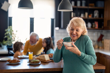 Happy carefree senior woman holding bowl with oatmeal muesli standing in the kitchen having breakfast at home. - obrazy, fototapety, plakaty