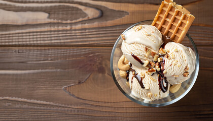 Glass bowl of ice cream dessert with chocolate, nuts and waffle, from above close-up on wooden background with copy space