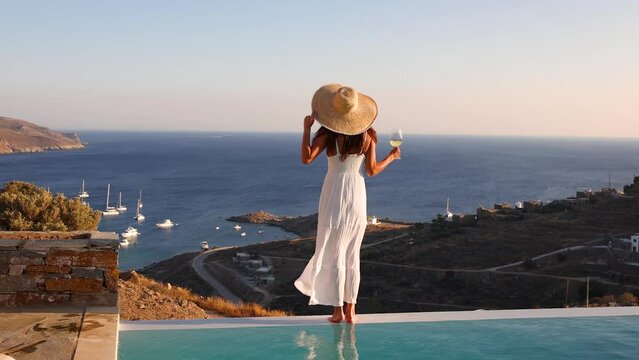 Attractive holiday woman in a white dress at the swimming pool enjoys the sunset over the mediterranean sea with a glass of wine