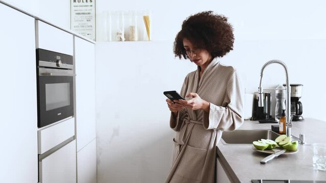 Positive african woman in glasses and housecoat texting on mobile in kitchen at home