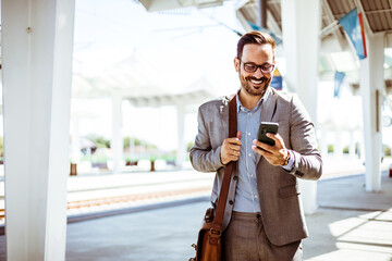 Photo of a smiling businessman at train station. Businessman using mobile phone at train station....