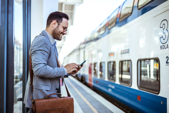 Handsome Young Man On Business Trip Walking With His Luggage And Talking On Cellphone At Train Station. Travelling Businessman Making Phone Call. Photo Of A Smiling Businessman At Train Station