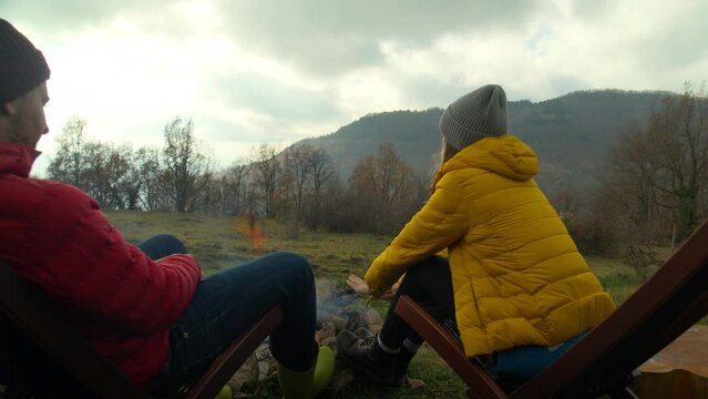 Zoom out shot of couple of young travelers rest by the fire, surrounded by autumn nature and mountains. Sunset at camping or natural hotel. Destination for an epic outdoor getaway.
