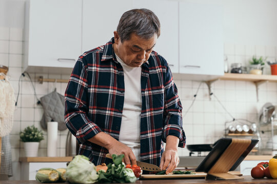 The Old Asian Man In The Kitchen, Using A Digital Tablet Pad To Learn Cooking Techniques, Chopping Vegetables On The Board With Ingredients In The Home Kitchen.