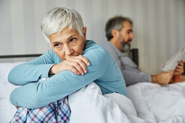 Portrait of unhappy senior woman or couple during or after arguing while sitting on bed, married elderly couple discussing in bed, or a health problem, mature woman being in pain