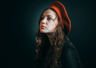 Portrait of a sad young woman in an orange beret and long curly hair on a dark background. - 622578994