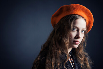 Portrait of a sad young woman in an orange beret and long curly hair on a dark background. - 622579121