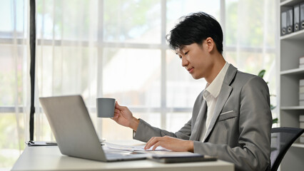 A smart Asian male boss reading business documents and sipping coffee at his desk.
