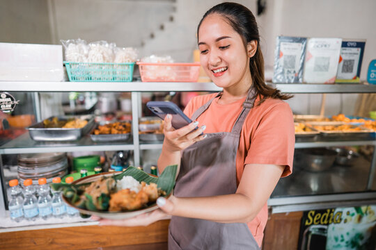 beautiful Asian girl in apron takes photo of pecel on plate using cell phone at local restaurant