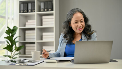 A happy Asian senior businesswoman looking at her laptop screen and working at her desk.