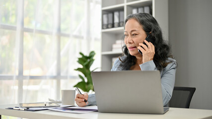 A professional Asian senior businesswoman is on the phone with her client at her desk
