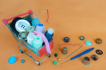 A shop cart with threads, sewing accessories and buttons. The background is made of light orange fabric.