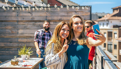 Portrait of two smiling female friends taking a selfie with phone in rooftop on a summer party while their colleagues laughing on background