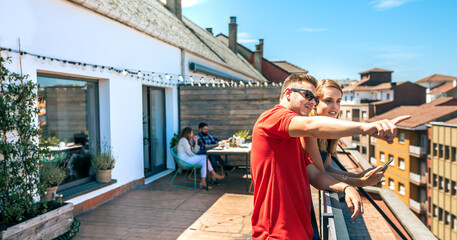 Smiling young man with sunglasses and blonde woman looking to the city from rooftop with their friends resting sitting on background