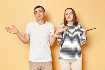 Uncertain puzzled young couple wife and husband wearing casual attires standing isolated over beige background shrugging shoulders don't know what to say.