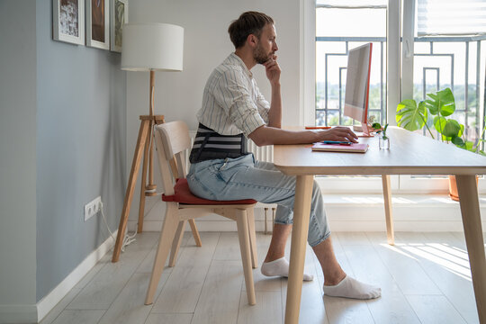Focused male freelancer wearing lumbar brace sitting at table using computer, young man use orthopedic corset belt while working remotely with herniated disc. Work from home and back pain 