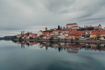 Obraz premium A panoramic view of Ptuj, Slovenia, reveals its charming old town, majestic castle, and a meandering river under a cloudy sky.